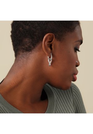 Porträt einer Person mit kurzen lockigen Haaren, die eine strukturierte silberne Knotenohrstecker und einen gerippten grau-grünen Pullover trägt, vor einem neutralen Hintergrund.