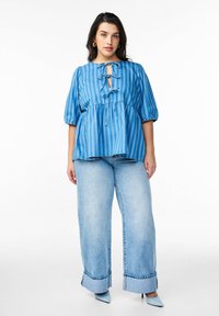 Modra prugasta bluza z naborki in vezavo spredaj, v kombinaciji z širokimi svetlimi jeans hlačami in modrimi špicastimi petami.