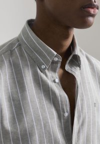 Camisa de lino gris con rayas verticales blancas, cuello de botón y dos botones visibles en el cuello. Presenta un logo sutil cerca del dobladillo.