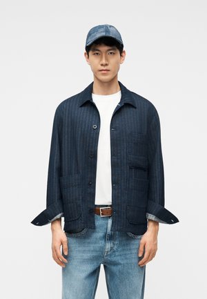 Jeune homme portant une veste rayée bleue par-dessus une chemise blanche, un jean bleu clair, une ceinture marron et une casquette bleue à motifs, debout devant un fond blanc.