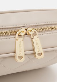 Light beige quilted leather bag with prominent gold zippers and hardware. Features embossed text on zipper pulls and textured trim.
