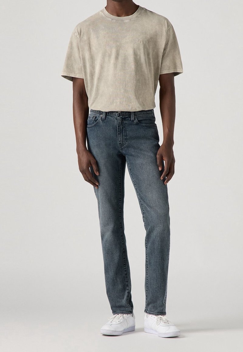 Homme portant un t-shirt beige délavé à manches courtes, un jean bleu foncé et des baskets blanches, debout devant un fond clair uni.