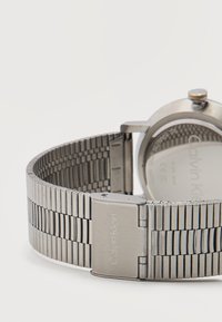 Ασημένιο μεταλλικό ρολόι Calvin Klein με συνδεδεμένο βραχιόλι και κούμπωμα, που εμφανίζει την πίσω όψη του προσώπου και του λουριού του ρολογιού σε λευκό φόντο.