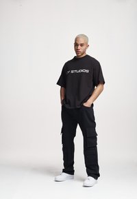 T-shirt noir surdimensionné avec le texte blanc "BY STUDIOS", associé à un pantalon cargo noir avec plusieurs poches et des baskets blanches.