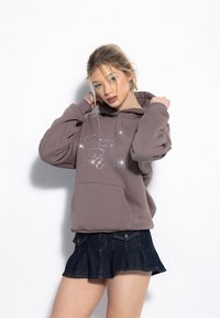 Brown hoodie with sparkling silver text "Good Friends," featuring a front pocket and loose-fit sleeves, paired with a denim skirt.