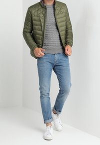 Giacca trapuntata verde oliva, maglione grigio testurizzato, jeans azzurri chiari e sneakers bianche su una persona in piedi contro uno sfondo chiaro.