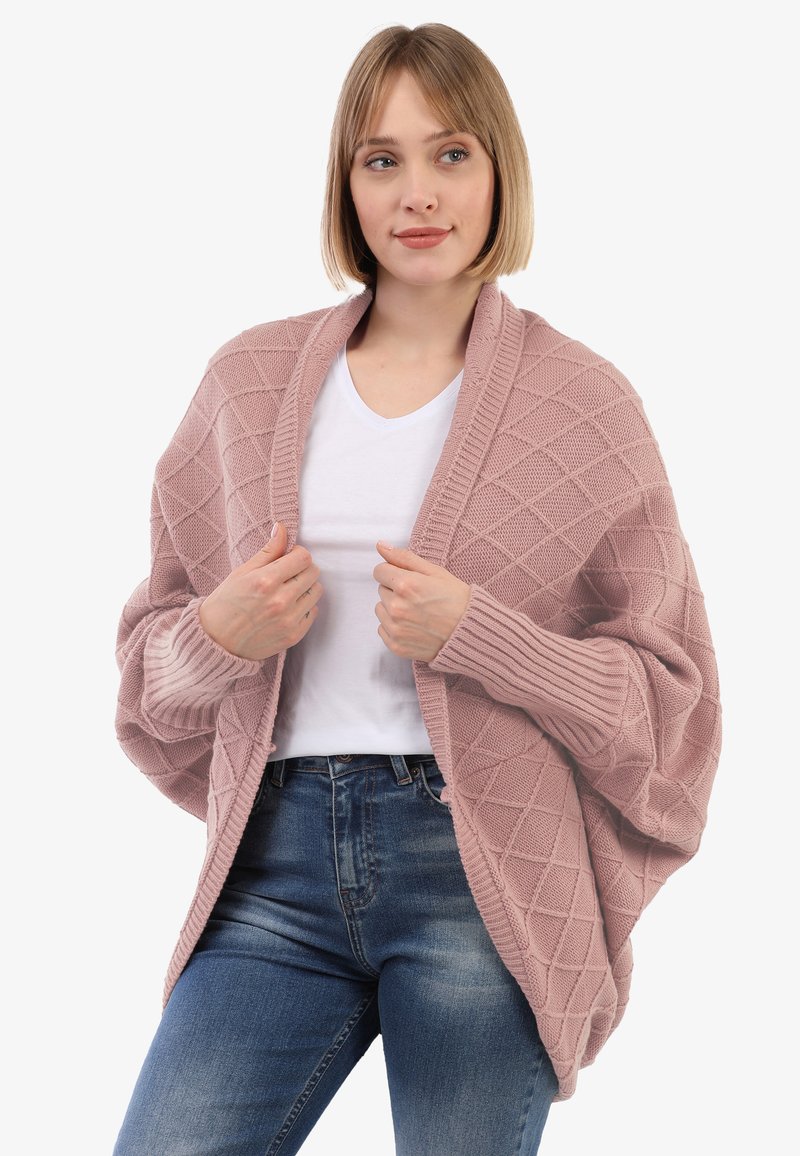 Cardigan lavorato a maglia rosa chiaro con motivo a diamante, apertura frontale e polsini a costine. Indossato sopra una camicia bianca e abbinato a jeans blu.