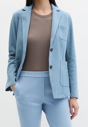 Person iført en lyseblå blazer og matchende bukser med en brun ribbet top, stående med den ene hånd i lommen mod en ensfarvet baggrund.