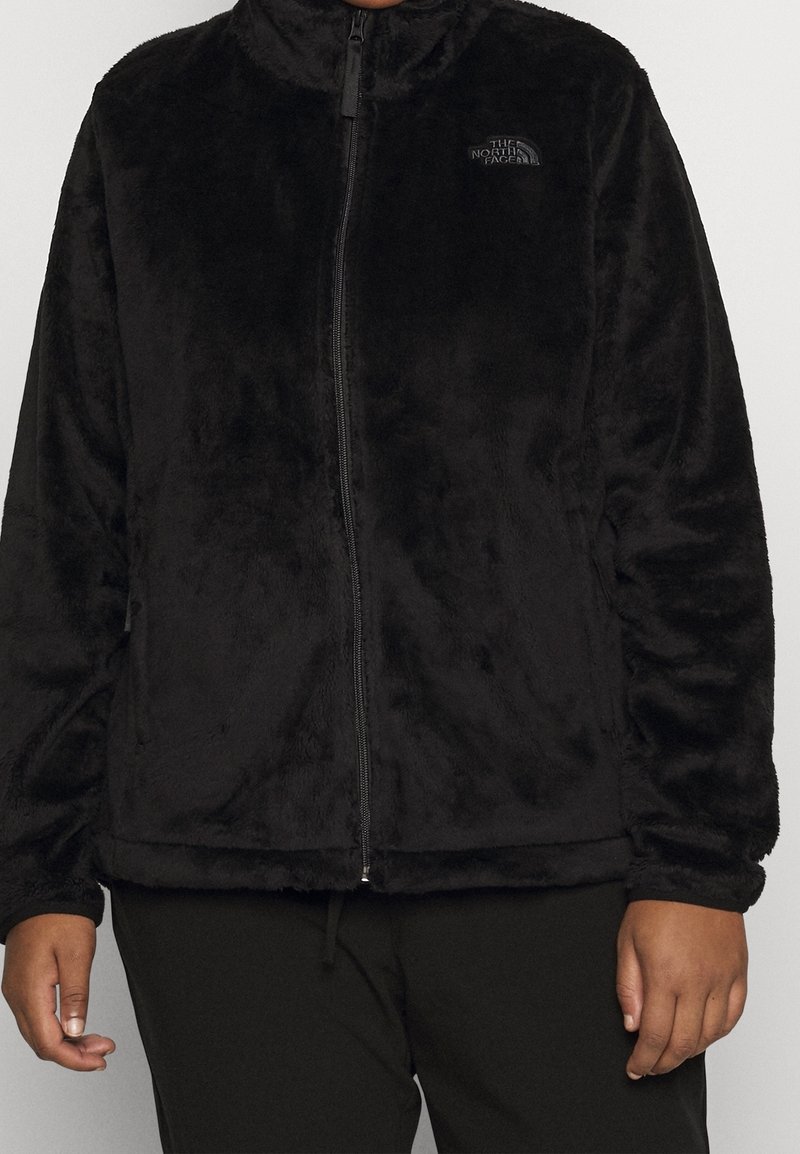 Giacca nera in pelliccia sintetica con zip e collo alto indossata sopra pantaloni neri, con un logo discreto sul lato sinistro del petto.
