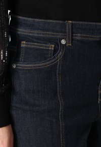 Σκούρο μπλε τζην παντελόνι με πορτοκαλί ραφές, διαθέτοντας μπροστινή τσέπη και μεταλλικό κούμπωμα. Το ύφασμα φαίνεται σταθερό με λεία υφή.