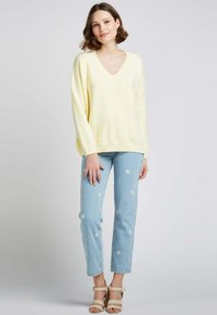 Pull oversized jaune clair avec un col en V, associé à un jean brodé bleu clair avec des motifs floraux et des sandales compensées beiges.