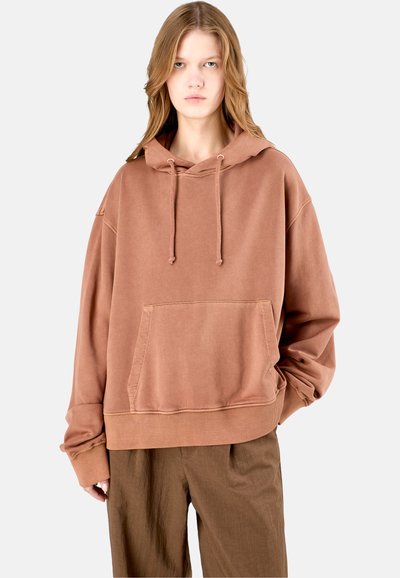 Bruine oversized hoodie met een voorzak, een capuchon met trekkoord en een ribbelrand. De stof lijkt zacht met een subtiele wassing. Gecombineerd met bruine broek.