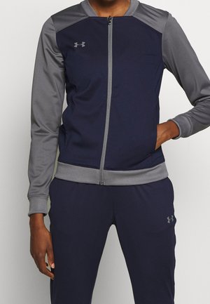 Mann trägt eine marineblaue und graue Sportjacke mit Reißverschluss und passende marineblaue Hose, mit der Hand in der Jackentasche.