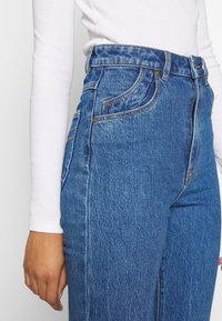 Džíny s vysokým pasem v modré denimové barvě s vybledlým vzorem, obsahující detaily šití, dvě přední kapsy a klasické zapínání na knoflík.