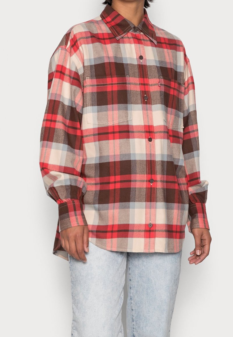 Chemise en flanelle à carreaux rouges et bruns avec un design boutonné, deux poches poitrine, manches longues et une coupe décontractée. Texture douce.