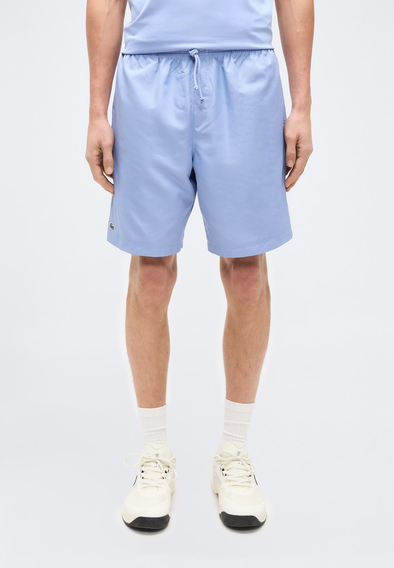 Άνδρας με ανοιχτά γαλάζια σορτς, λευκά αθλητικά παπούτσια και λευκές κάλτσες, στέκεται μπροστά σε ένα απλό φωτεινό φόντο.