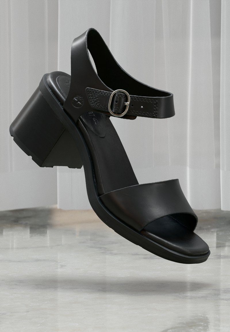 Μαύρο δερμάτινο πέδιλο με ανοιχτό δάχτυλο, τετράγωνο τακούνι και ρυθμιζόμενο λουρί στον αστράγαλο με ασημί πόρπη, τοποθετημένο σε μαρμάρινο πάτωμα.