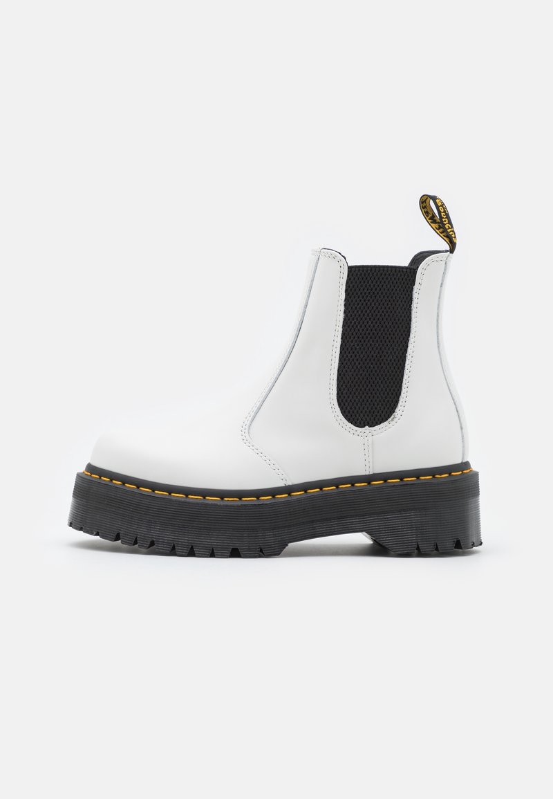Bottine Chelsea en cuir blanc avec panneau latéral élastique noir, semelle noire épaisse, couture jaune et languette de tirage marquée sur fond neutre.