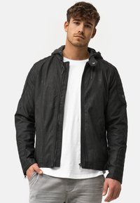 Schwarze Kunstlederjacke mit durchgehendem Reißverschluss, Kapuze und gerippten Akzenten an den Schultern, getragen über einem weißen T-Shirt, kombiniert mit hellgrauen Hosen.