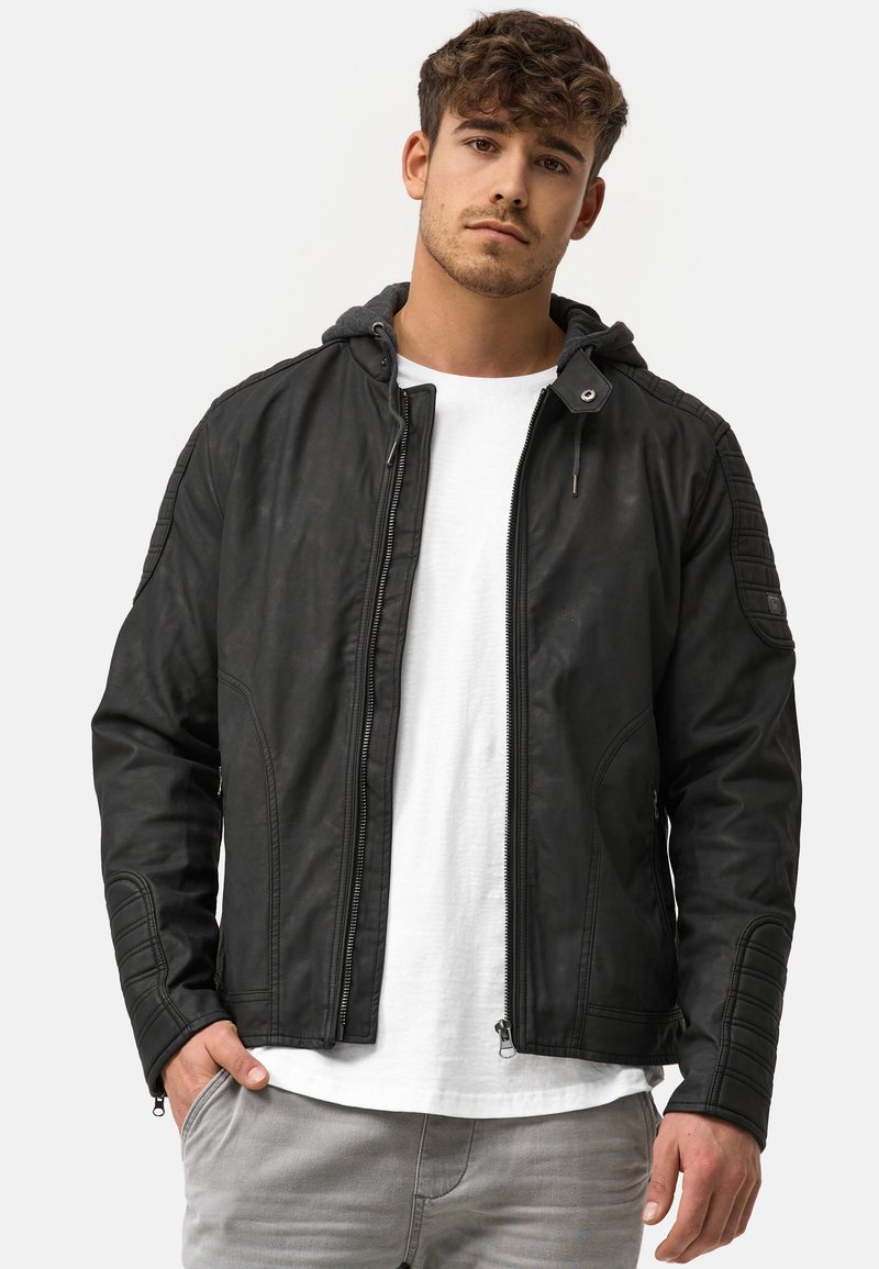 Schwarze Kunstlederjacke mit durchgehendem Reißverschluss, Kapuze und gerippten Akzenten an den Schultern, getragen über einem weißen T-Shirt, kombiniert mit hellgrauen Hosen.