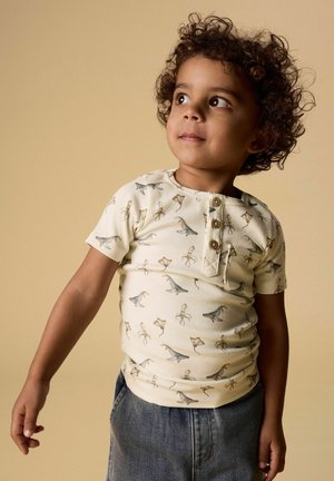 Małe dziecko o kręconych włosach, ubrane w koszulkę z krótkim rękawem w wzór dinozaurów oraz dżinsowe spodnie, patrzące lekko w górę i na bok.