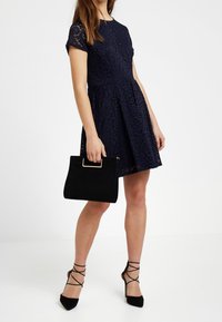 Námořnické modré krajkové šaty s krátkým rukávem a rozšířenou sukní, doplněné o černou semišovou kabelku a černé sandály na podpatku.