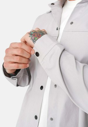 Camisa gris claro de botones con herrajes negros, que presenta un cuello estructurado y un puño ajustado con botones, llevada sobre una camiseta interior blanca.