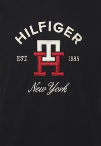 Černé tričko s červeným a bílým vyšívaným logem s nápisem "HILFIGER Est. 1985 New York" s význačným písmem a designovými prvky.
