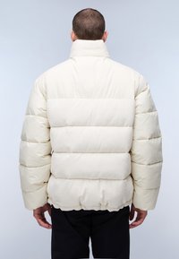 Crème pufferjack met een hoge kraag, voorzien van gequilte secties en ribbelstof op het middelste gedeelte. Elastische zoom en manchetten.