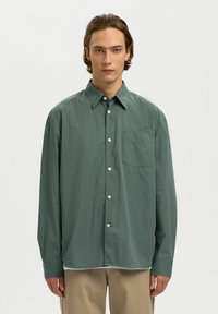 Homme portant une chemise boutonnée ample vert foncé et un pantalon beige, faisant face à l'avant sur un fond clair uni.