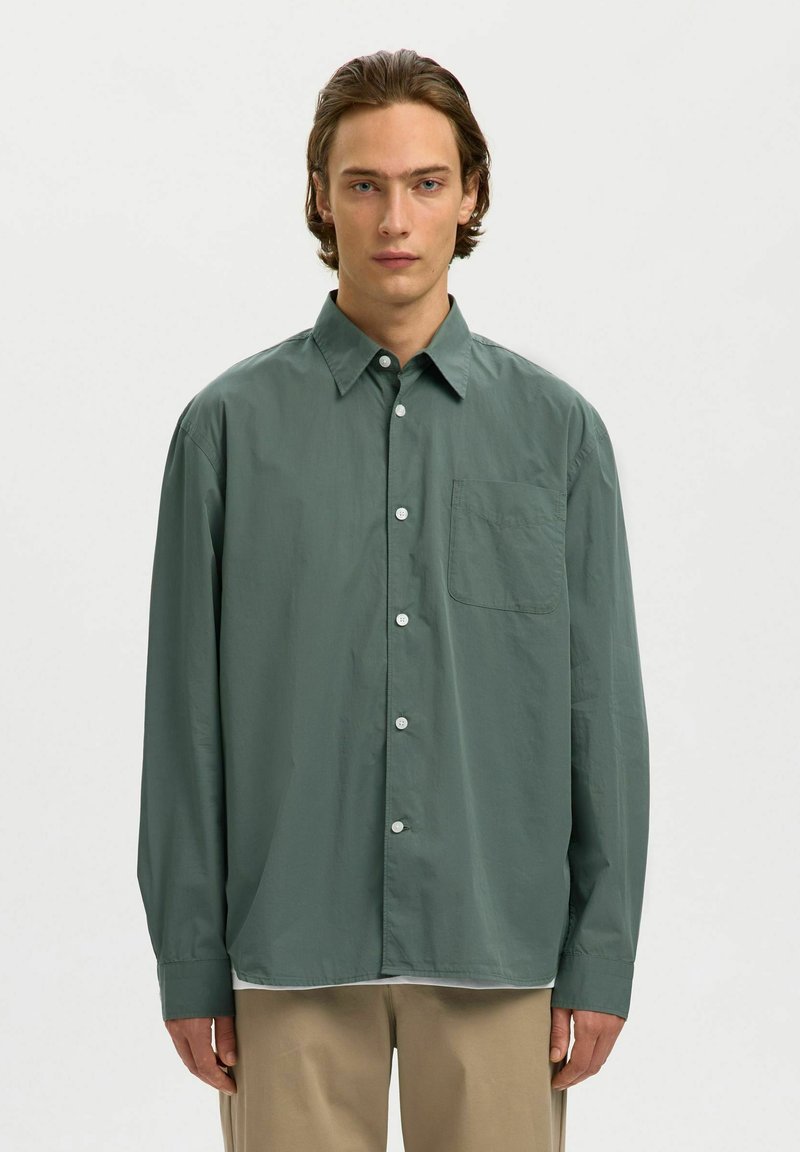 Homme portant une chemise boutonnée ample vert foncé et un pantalon beige, faisant face à l'avant sur un fond clair uni.