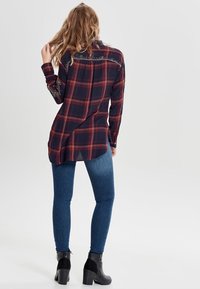 Long-sleeve plaid shirt in navy, red, and white with decorative stitching on shoulders, paired with fitted blue jeans and black heeled ankle boots.