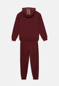 Maroon Nike dukserica s kontrastnim logotipom na kapuljači, uparen sa odgovarajućim maroon trenirkama. Mekani materijal i rebrasti manžeti.