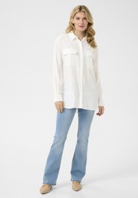 Camicia bianca a maniche lunghe con bottoni, dotata di due tasche sul petto, abbinata a jeans svasati blu chiaro e scarpe beige, il tutto su uno sfondo neutro.