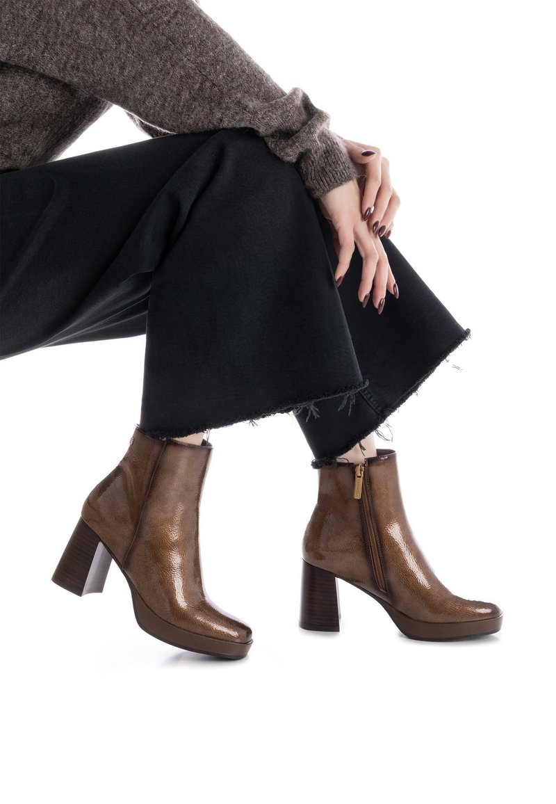 Botas de cuero marrón hasta el tobillo con un acabado brillante, que cuentan con un zipper lateral y un tacón grueso. El diseño incluye una puntera redondeada y detalles texturizados.