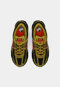 Pár čiernych a olivových tenisiek Nike Vomero 5 s oranžovými akcentmi a žltými šnúrkami zobrazený zhora na bielom pozadí.