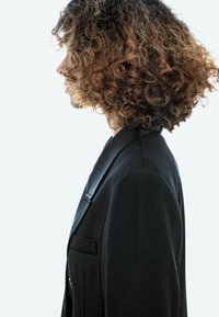 Schwarzer Blazer mit Satinrevers, taillierter Form, strukturierten Schultern und seitlichen Taschen. Lockiges Haar ist am Profil des Modells sichtbar.