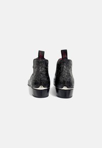 Botines negros hasta el tobillo con patrones florales en relieve, tacones negros brillantes y tiradores rojos en la parte posterior. Parte superior de cuero texturizado.