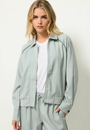 Blonde vrouw draagt een lichtgroene fijn gestreepte jas met rits en bijpassende broek met drawstrings over een wit t-shirt, poseert met één hand in de zak.