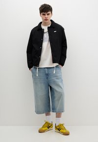Črna lahka jakna, bela grafična majica, svetlo modri široki jeans kratki hlač, in rumeni športni čevlji s črnimi črtami. Bela ozadje.