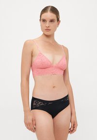 Kvinne iført rosa blonde bralette med tynne stropper og svarte hipster-truser med blondekant, stående mot hvit bakgrunn, og ser litt mot venstre.