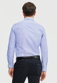 Niebieska i biała w paski koszula z długim rękawem, wykonana z bawełnianego materiału, z guzikami przy mankietach, dopasowany krój, noszona z ciemnymi spodniami, widok z tyłu.