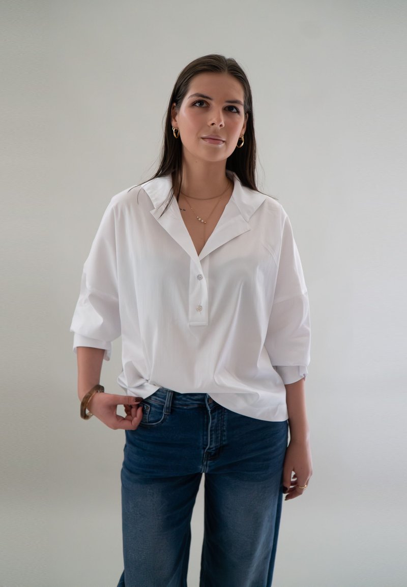 Vrouw met lang zwart haar, gekleed in een losse witte blouse met knopen, blauwe spijkerbroek, gouden oorringen en gelaagde kettingen, tegen een effen achtergrond.