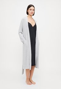 Light gray long cardigan with open front, ribbed cuffs, side pockets, worn over a black slip dress. Soft knit fabric, knee-length design.