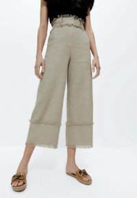 Světle béžové široké kalhoty z texturované látky, s třásněmi na lemování a vysokým pasem. V kombinaci s hnědými espadrilami.