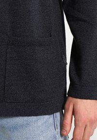 Ciemnoszary sweter z fakturowanej tkaniny, z przednią kieszenią i widoczną metką marki po boku. Rękawy długie i dopasowane.