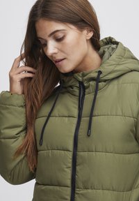 Zelená péřová bunda s kapucí, která má prošívaný design, černý zip a šňůrky na stažení. Na povrchu je hladká textura a viditelné teplé izolační vlastnosti.