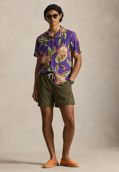 Άνδρας με μωβ κοντομάνικο πουκάμισο με λουλούδια, λαδί σορτς, πορτοκαλί παπούτσια χωρίς κορδόνια, γυαλιά ηλίου και ρολόι χειρός, στέκεται μπροστά σε ουδέτερο φόντο.