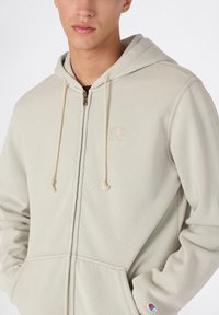 Sudadera con cremallera de color beige claro, hecha de tejido texturizado, con capucha ajustable, bolsillos frontales y un pequeño logotipo bordado en el pecho.