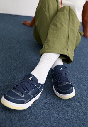 Personne assise sur un tapis bleu portant un pantalon vert, des chaussettes blanches et des baskets bleu marine avec des semelles blanches.